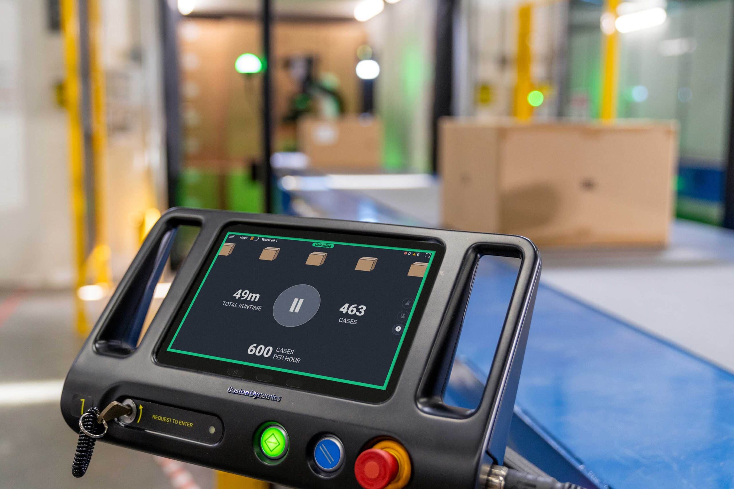 A console to operate a robot displays metrics on screen and has colorful buttons at the bottom. Behind it in soft focus, a robot unloads a container and boxes travel down a conveyor belt.